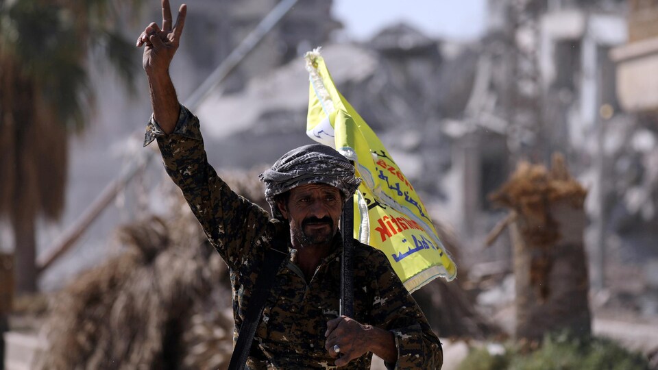 Un combattant des Forces démocratiques syriennes (FDS), qui marche dans la ville en ruine, tient un drapeau sur lequel des mots arabes sont écrits et fait un signe de paix avec sa main droite.