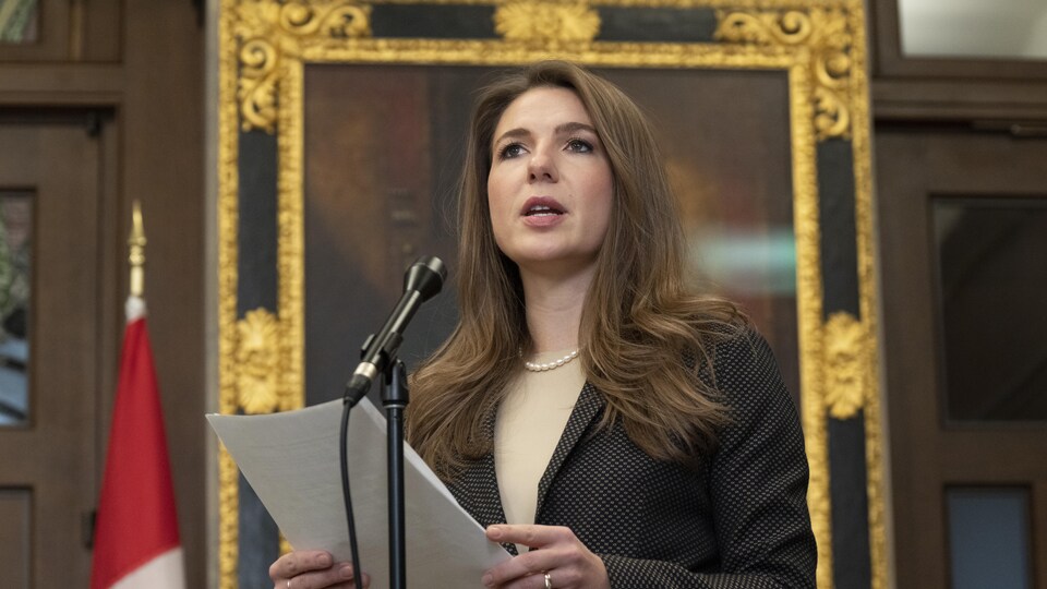 Raquel Dancho debout devant un micro sur pied, des documents à la main. En arrière-plan, il y a un cadre doré.