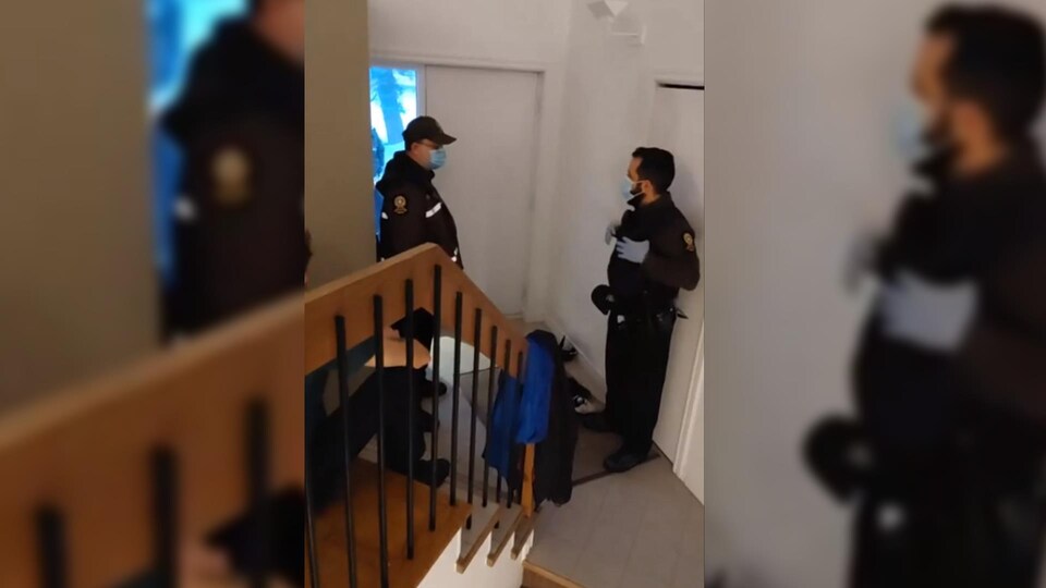 Deux policiers, debout dans l'entrée d'une maison, discutent. Ils portent un masque.