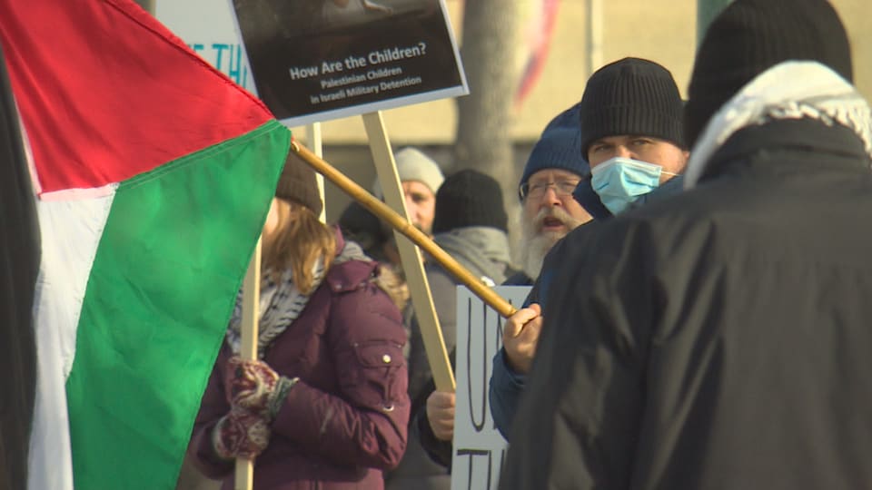 Des gens sont debout dehors en manteau et brandissent des pancartes et un homme tient un drapeau de la Palestine. 