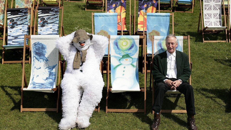 Vêtu d'un complet vert, l'illustrateur Raymond Briggs est assis sur une chaise pliante à côté d'une personne déguisée en bonhomme de neige qui se prélasse sur une chaise similaire.
