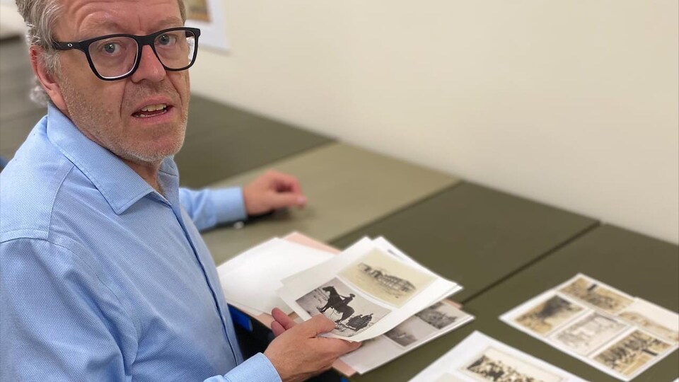 Raymond Frogner, assis, se tourne vers la caméra. Devant lui, des photos d'archives sont disposées sur un classeur.