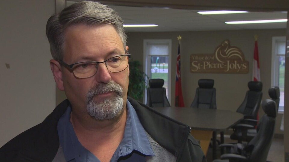 Le maire Raymond Ménard en gros plan, dans la salle du conseil de Saint-Pierre-Jolys. En arrière-plan, la table avec les chaises des conseillers et au mur, une plaque avec le nom du village.