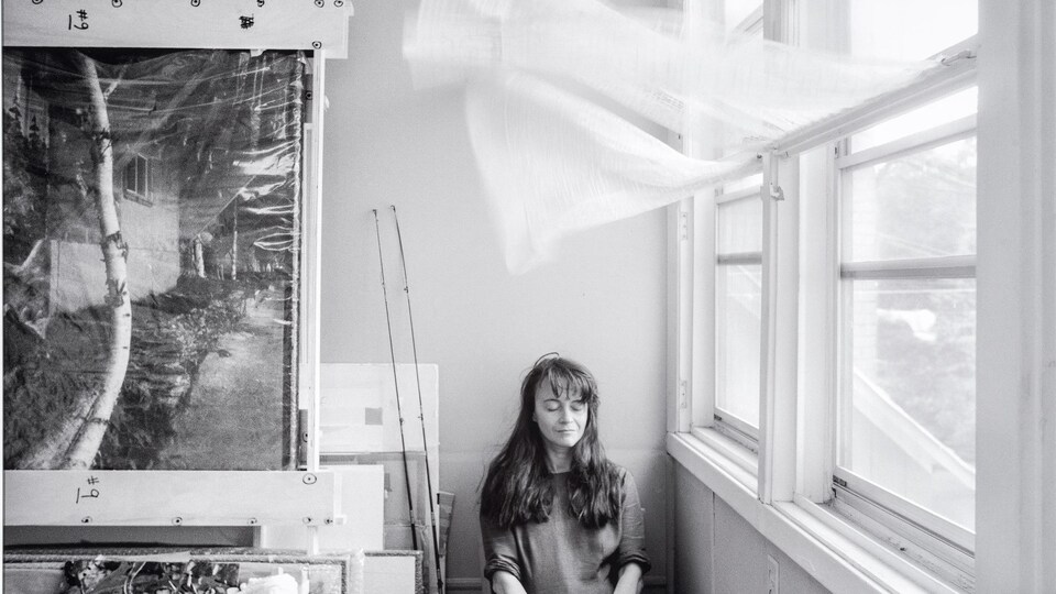 Une femme est assise devant une fenêtre à l'intérieur, un rideau est soulevé par le vent au-dessus d'elle et des tableaux sont à sa droite. La photo est en noir et blanc.