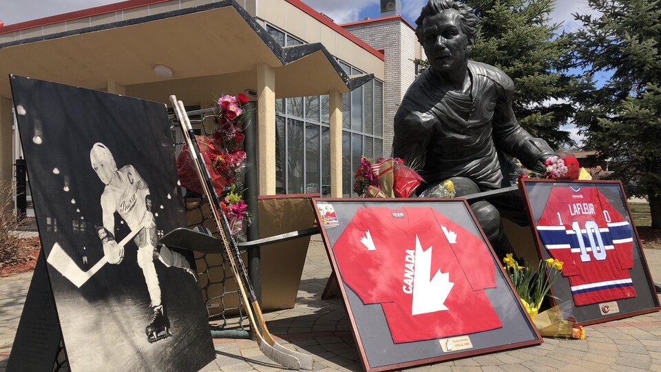 Des cadres, des bâtons de hockey et des fleurs devant une stature de Guy Lafleur.