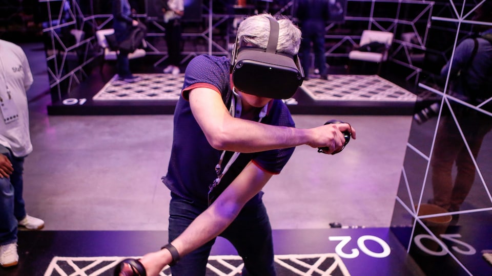 Une personne porte un casque de réalité virtuelle Oculus et tient deux manettes dans les mains. La personne est dans un centre où se tient une conférence de jeux vidéo.