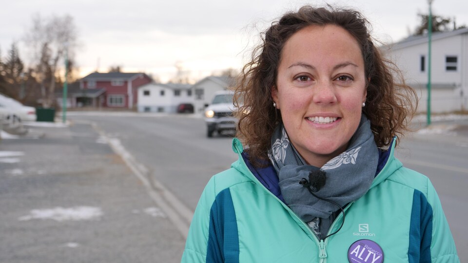 Rebecca Alty sourit pour la caméra devant une rue de Yellowknife. Elle porte un écusson de sa campagne.