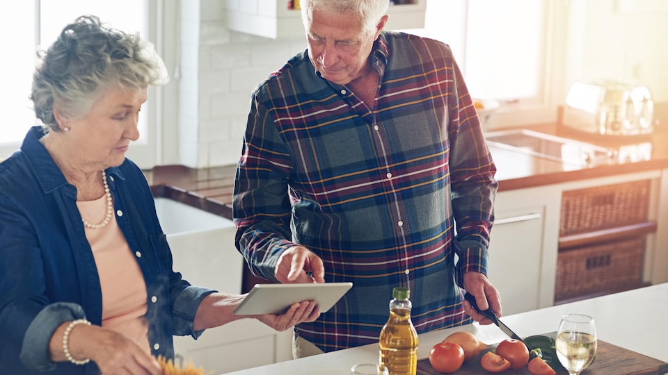 Un couple de personnes âgées prépare un repas en consultant une recette sur une tablette électronique.