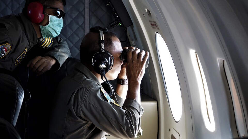 Des hommes portant des casques d'écoute regardent par le hublot d'un avion. Un des deux utilise des jumelles.