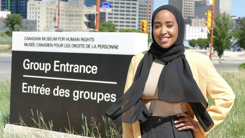 Une jeune femme est souriante. Elle se trouve devant l'entrée du Musée canadien pour les droits de la personne. 