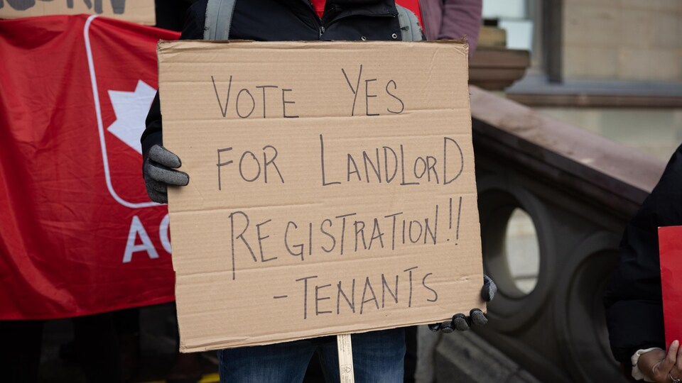 Plan rapproché d'une pancarte dans une manifestation. Sur l'affiche en carton, il est écrit : « Votez oui au registre des propriétaires ».