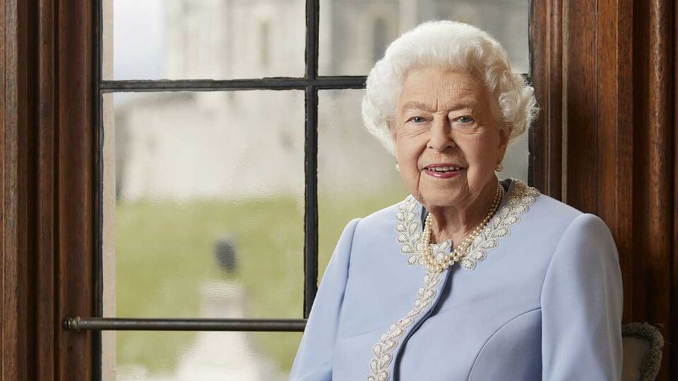 La reine Élisabeth II, assise près d'une fenêtre, vêtue d'habits bleus.
