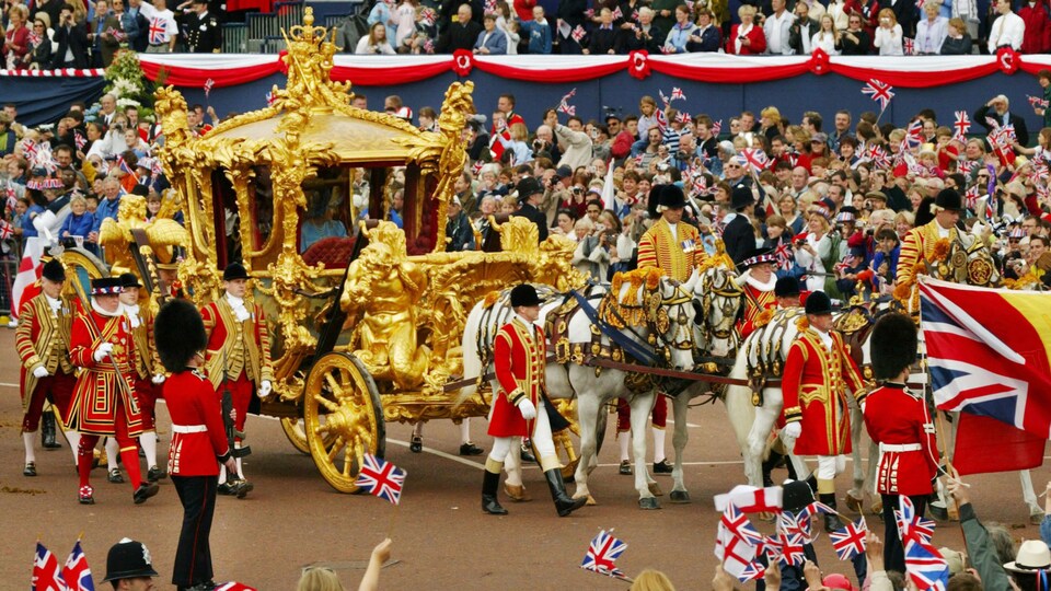 La reine est assise à l'intérieur d'un carrosse doré tiré par des chevaux qui circule dans une rue bordée de manifestants brandissant des drapeaux britanniques.
