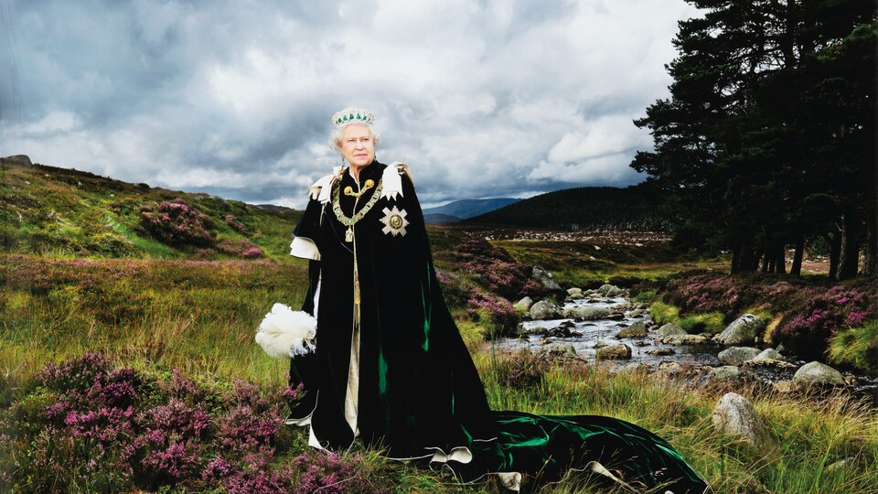 La reine Élisabeth II pose devant des collines verdoyantes, tenant un bouquet blanc dans sa main et regardant au loin.
