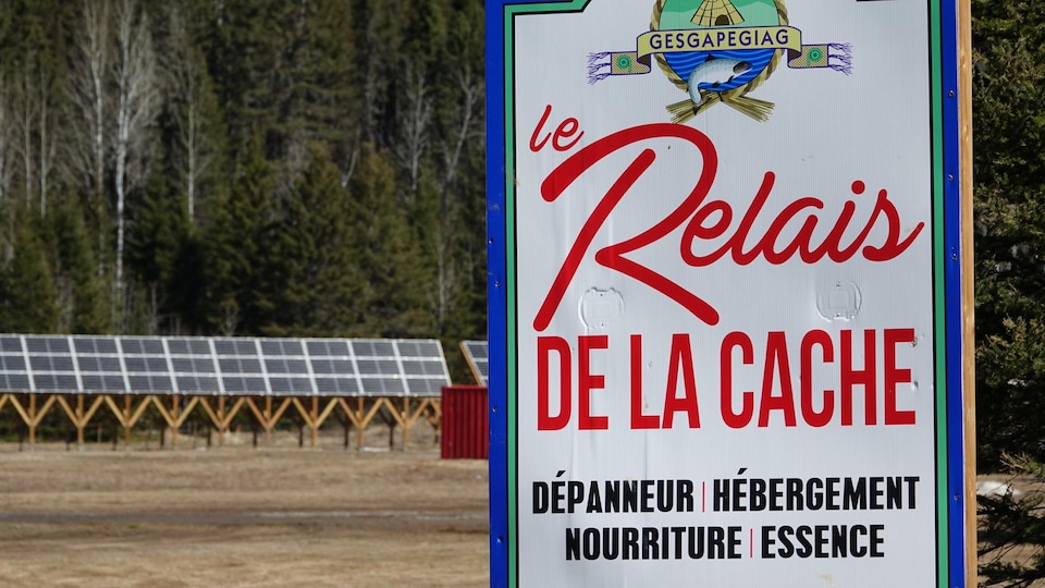 Une pancarte où il est écrit «Le Relais de la Cache: dépanneur, hébergement, nourriture, essence», avec des panneaux solaires en arrière-plan.