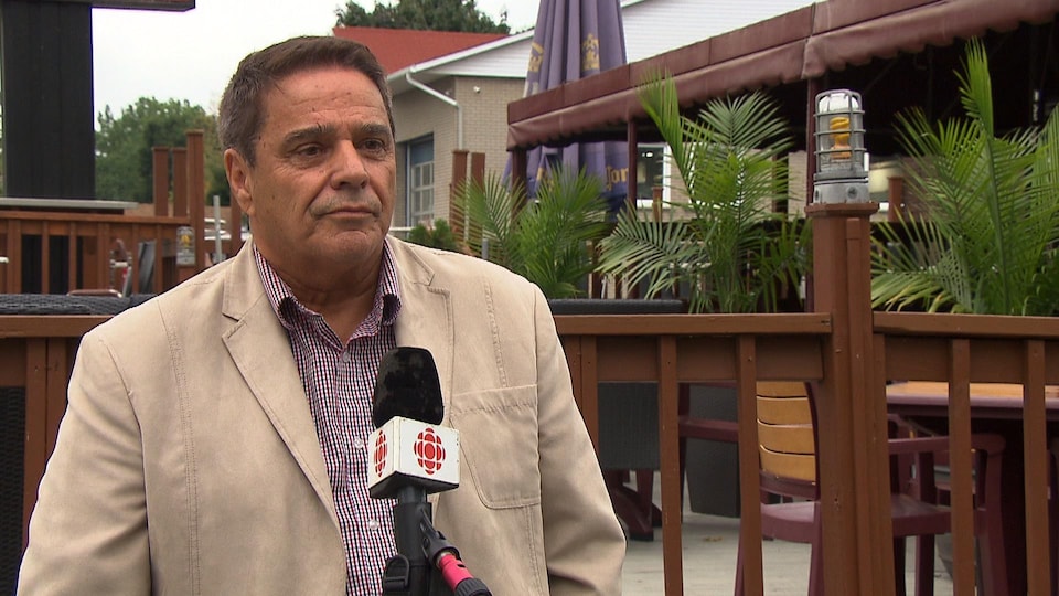Renaud Poulin porte un veston beige et une chemise à carreaux, il parle devant un micro, il est dehors devant la terrasse d'un restaurant.