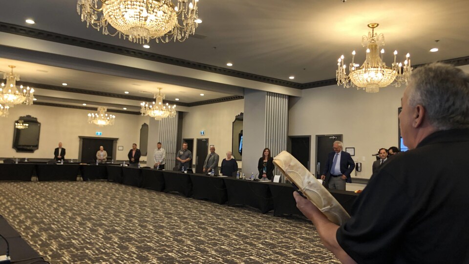 Un homme joue du tambour et plusieurs personnes sont debout autour d'une très grande table en rectangle, sous des chandeliers.
