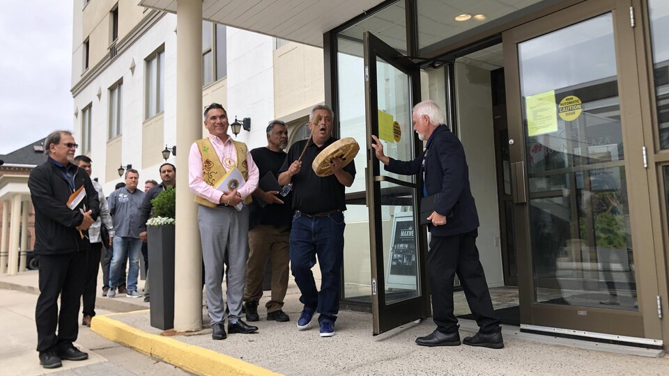 Un groupe de représentants des Premières Nations sur le perron d'un édifice. L'un d'entre eux frappe sur un tambour. Un homme leur ouvre la porte.