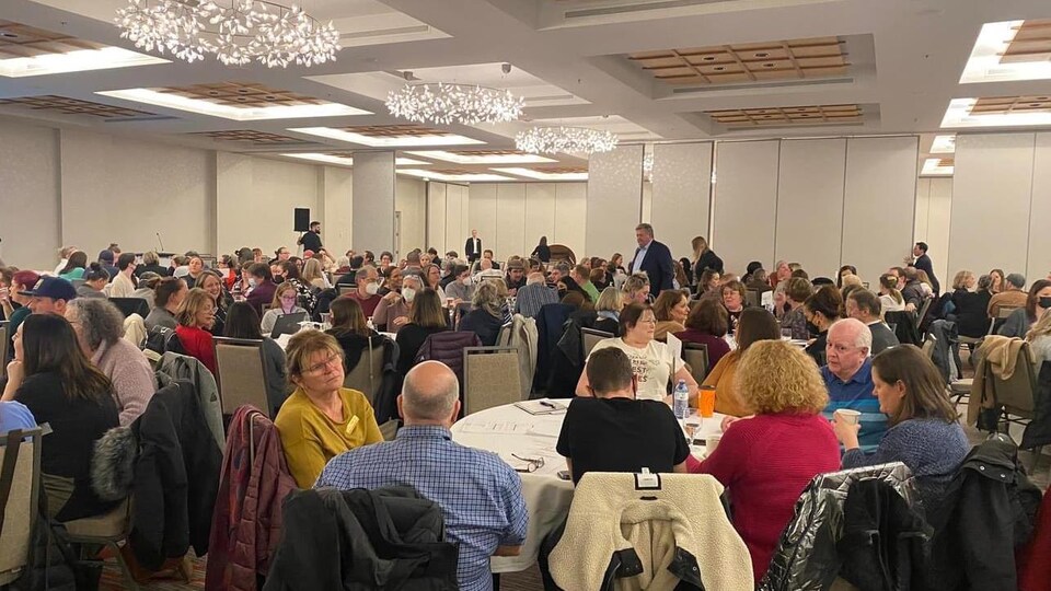 Des participants à une rencontre publique sont assis à des tables dans une salle comble.