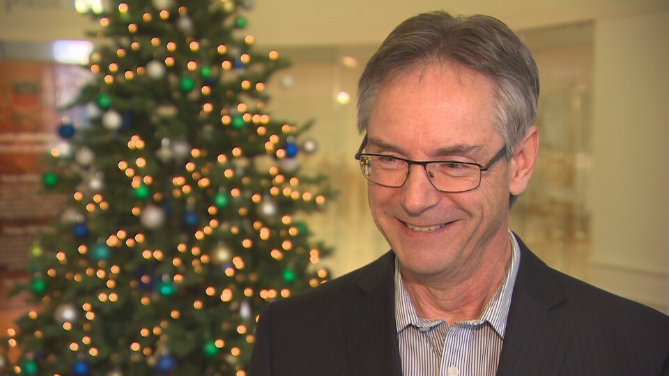 René Bouchard sourit. Il est photographié en gros plan avec un sapin de Noël en arrière-plan.