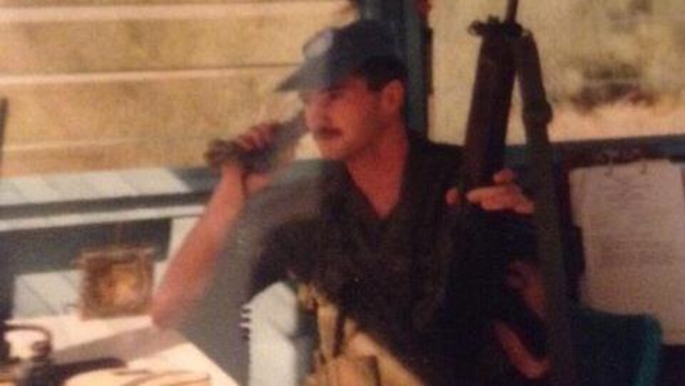 Un militaire coiffé d'une casquette tient un téléphone d'une main et une arme de l'autre.
