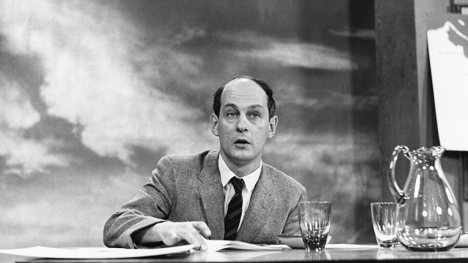 Dans un studio de télévision, assis à une table où reposent une carafe et deux verres, l'animateur René Lévesque s'apprête à tourner avec son index la feuille devant lui.