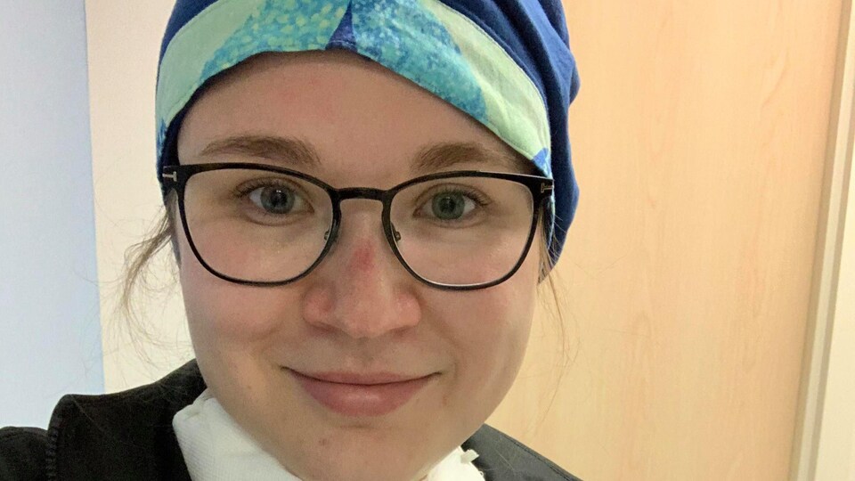 Égoportrait d'une jeune femme souriante. Elle porte des lunettes, blouse d'infirmière et un bonnet chirurgical