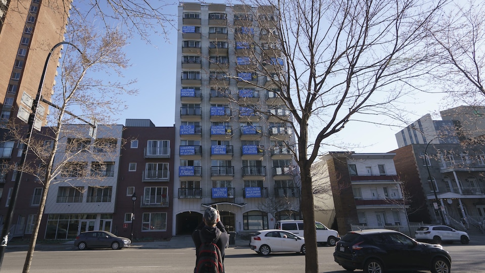 Une personne prend une photo d'un immeuble dont plusieurs balcons sont placardés par des pancartes bleues.