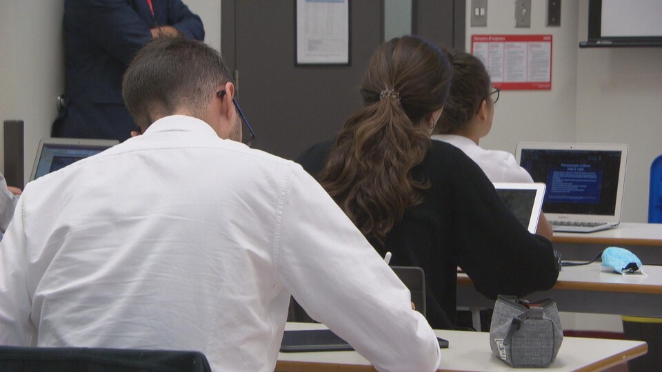 Des étudiants sont assis à des tables, ordinateurs devant eux. Ils regardent vers l'avant ou prennent des notes.