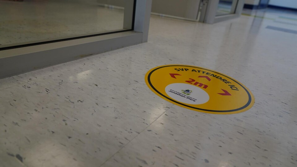 Un collant sur lequel il est écrit « SVP attendre ici » est visible dans un couloir.