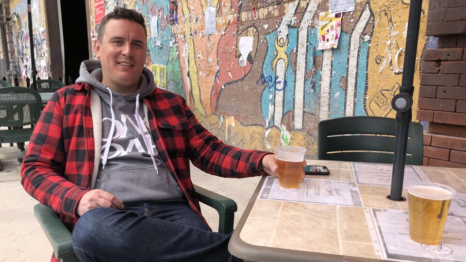 Un jeune homme souriant est assis à une table à l'extérieur, un verre de bière à la main.
