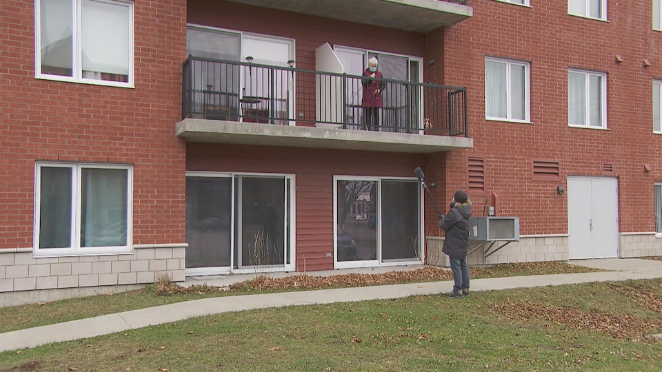 Un journaliste de Radio-Canada tend un micro accroché à une perche vers une résidente qui lui parle depuis son balcon. Tous les deux portent le masque.
