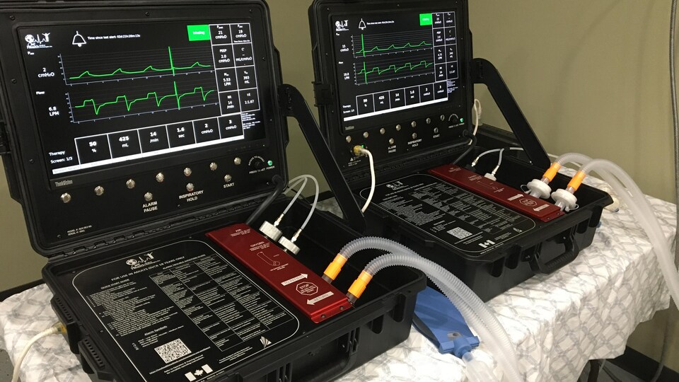 Deux respirateurs, côte à côte, similaires à une valise, ouverte, avec un écran et des boutons de contrôle dans la partie supérieure et un système de branchement dans la partie inférieure. 