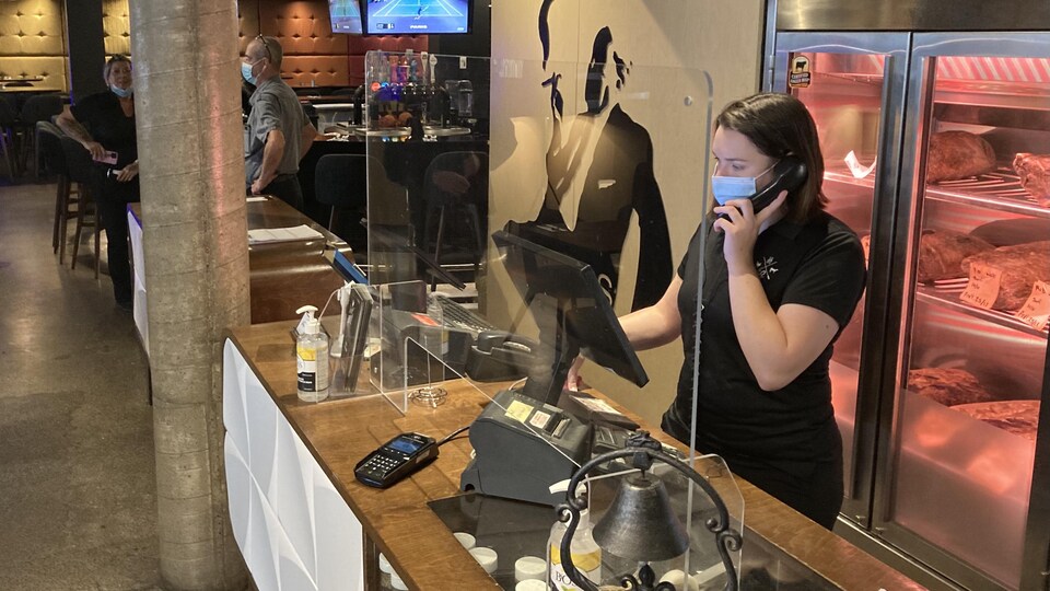 Une serveuse prend une réservation derrière un plexiglas à l'entrée d'un restaurant.