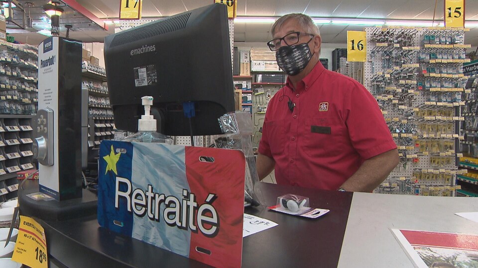 Une personne âgé est à son poste, devant une caisse, dans une quincaillerie. Une pancarte au bas de l'écran indique 'retraité'.