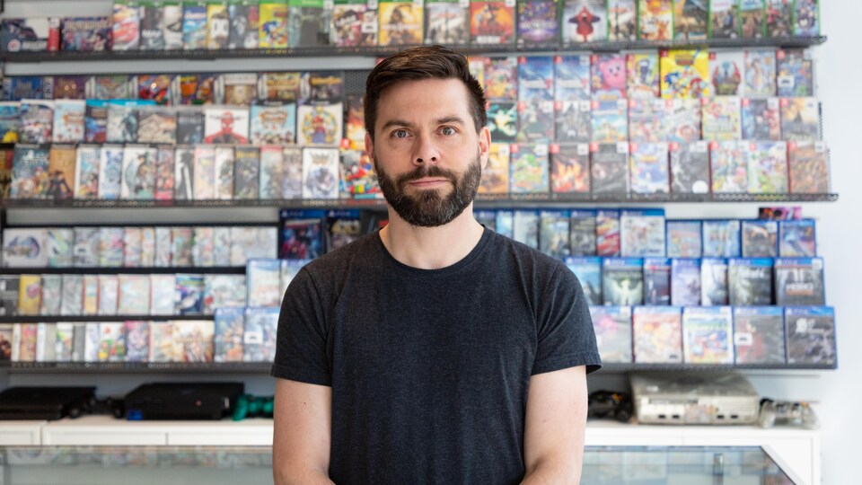 Une photo de Dominic Bourret, un homme dans la trentaine aux cheveux bruns courts portant une courte barbe, photographié devant un présentoir mural rempli d'emballages de jeux vidéo.