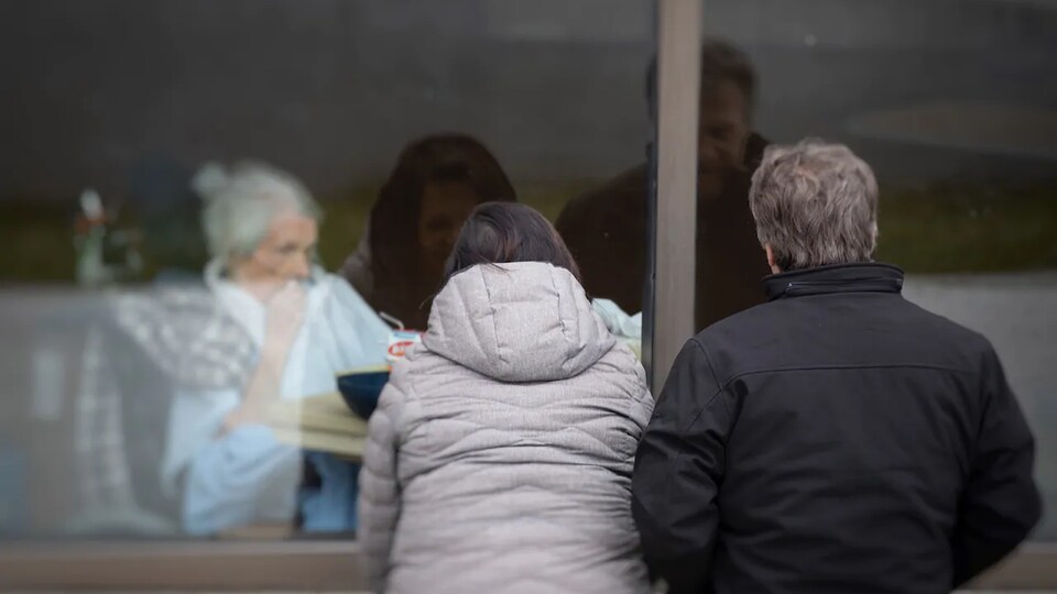 Un couple vu de dos regarde une aînée à travers une vitre.