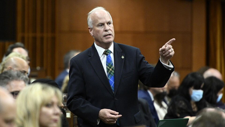 Le député est debout aux Communes. Il porte une cravate bleue et verte aux couleurs du tartan de la Nouvelle-Écosse. Il pointe vers sa gauche avec son index pendant qu'il parle.