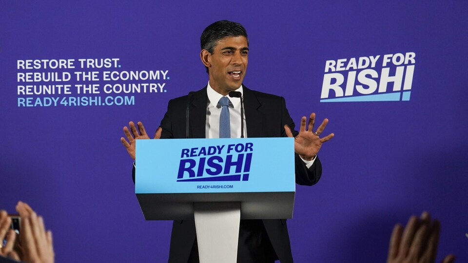 Un homme souriant, debout derrière un lutrin, devant un panneau illustrant un slogan politique.