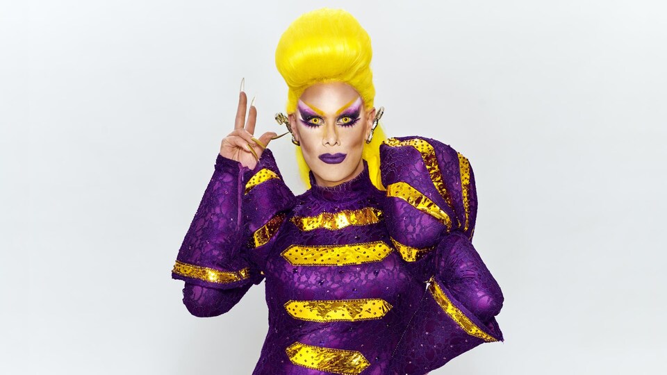 Rita Baga porte une perruque jaune ainsi qu'un costume mauve et doré.
