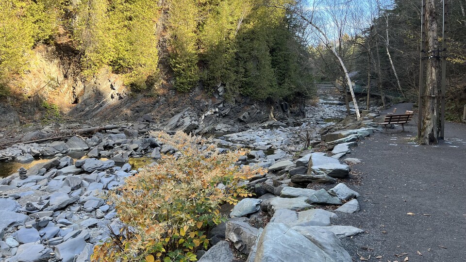 Un passage piétonnier en gravier borde une rivière dont le niveau est très bas. On voit essentiellement de gros rochers.