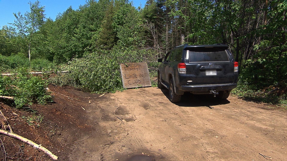 Un véhicule est stationné devant un amas d'arbres qui obstrue le chemin de terre. Une pancarte en bois indique : «accès fermé, limite du club».