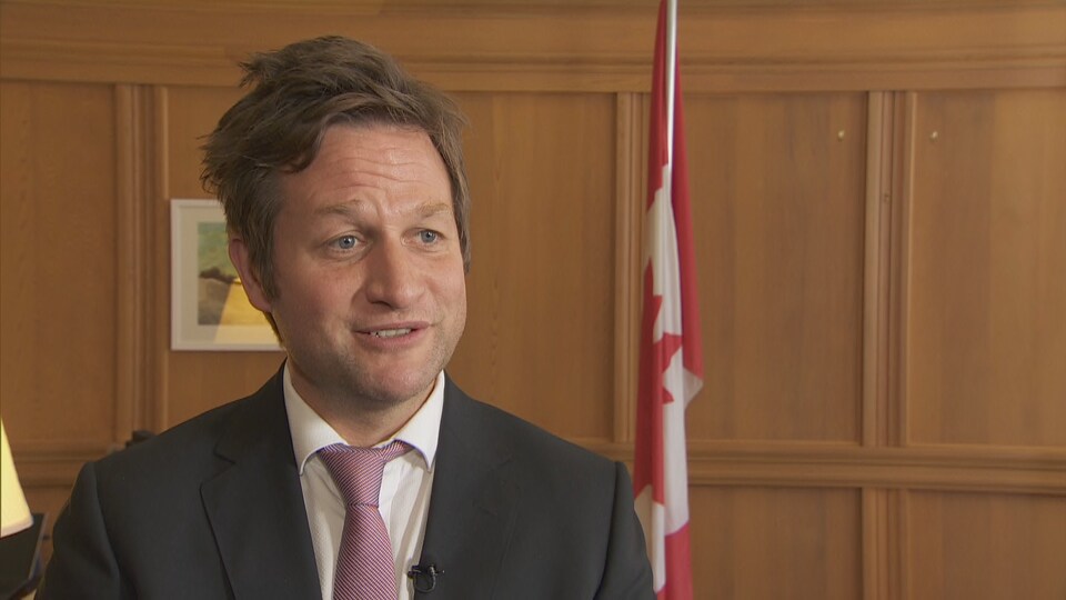 Le ministre de l'Éducation en entrevue à la caméra. Un drapeau du Canada figure derrière lui. 