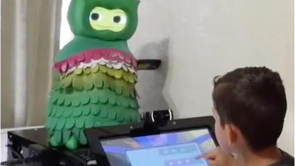 Un enfant interagit avec une tablette attachée à un robot qui ressemble à un hibou avec un visage humain.