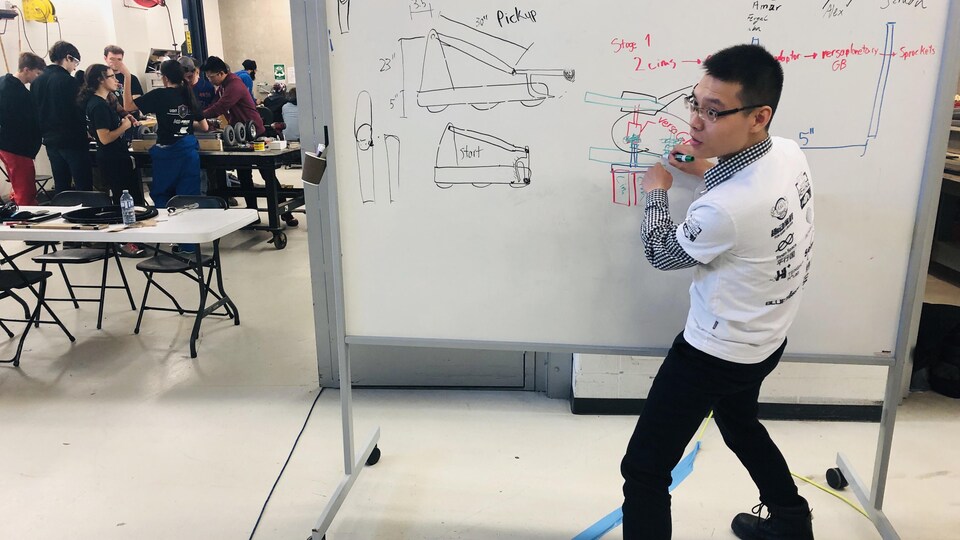 Dans le fond, à gauche, des étudiants montent le robot, au premier plan, à droite, un étudiant dessine sur un tableau blanc le plan du bras mécanique du robot.