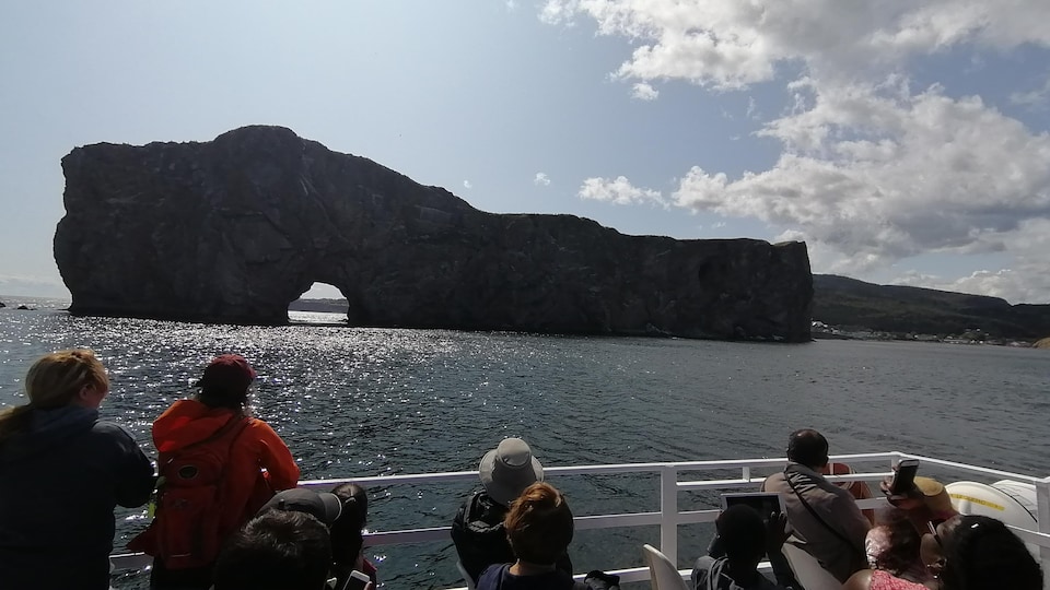 Dans un bateau de croisière, des touristes tiennent des cellulaires qu'ils pointent en direction du rocher Percé, au large.