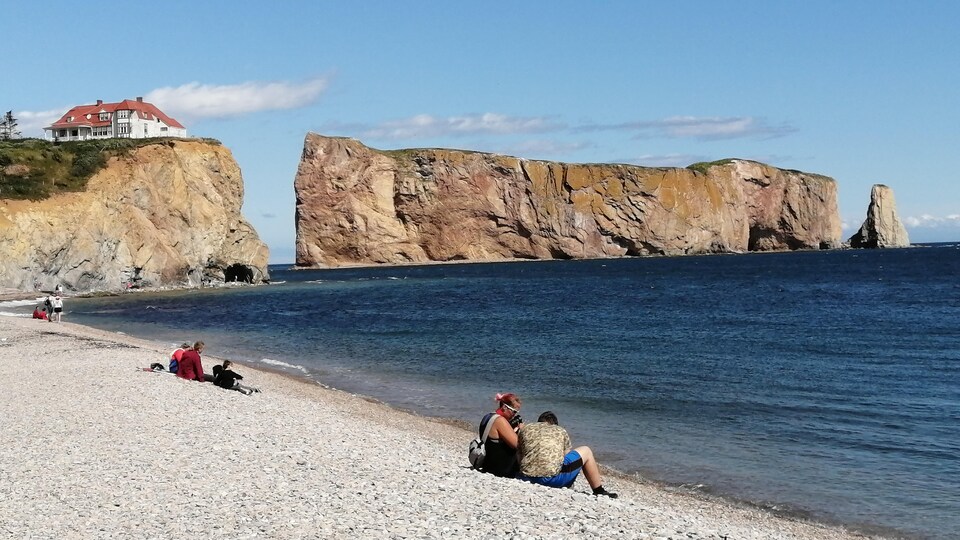 Des touristes sont assis sur une plage de galets. À l'arrière-plan, on voit le rocher Percé et un bâtiment patrimonial sur une colline.