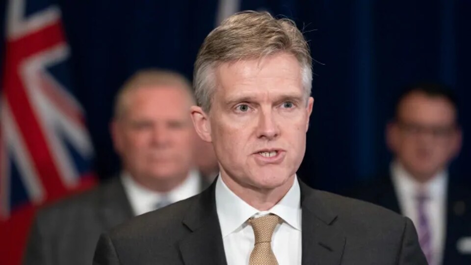 Un homme en complet parle avec deux personnes en arrière-plan.