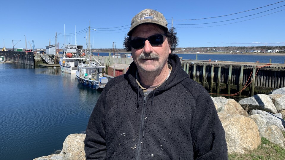 Le pêcheur porte une casquette et des lunettes de soleil et observe les bateaux amarrés au quai de Meteghan.