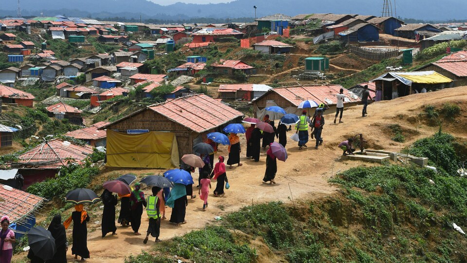 Des personnes marchent sur un petit chemin de terre près de milliers d'habitations. Les personnes ont un parapluie ouvert au-dessus de leur tête.